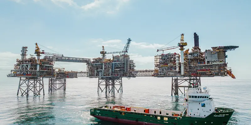 A large offshore oil rig with complex machinery stands over calm ocean waters under a partly cloudy sky. A green supply vessel is visible in the foreground.