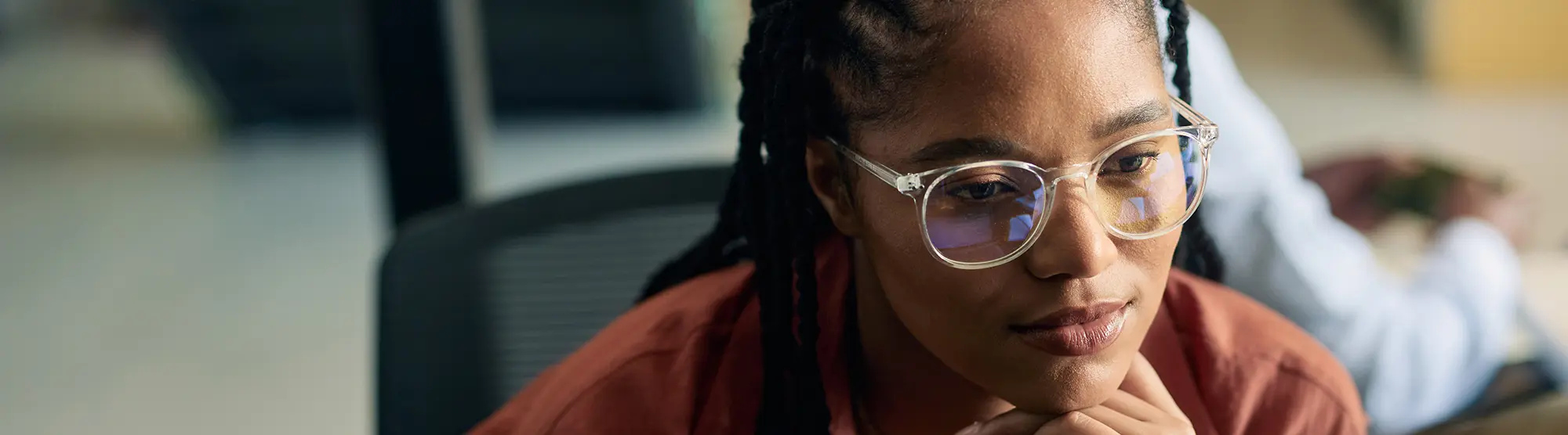 A focused individual with glasses, wearing a rust coloured top, looks intently at a computer screen. The background is softly blurred, conveying concentration.