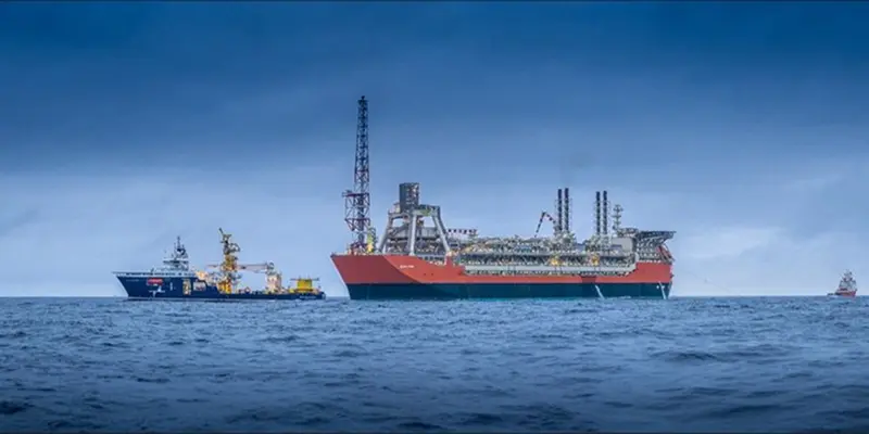 Large red and white offshore oil platform vessel with cranes floats on calm sea under a cloudy blue sky. Support ships visible nearby